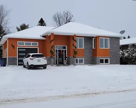 A raised bungalow with warm orange board-and-batten accents, an attached garage and stone base, in winter