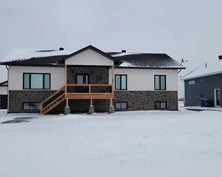 A white board-and-batten home with a stone base, cedar deck and an attached shop, in a snowy rural setting